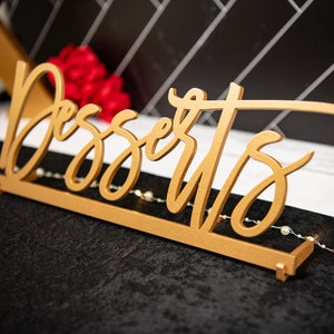 May include: A gold-coloured wooden sign with the word "Desserts" written in a decorative script font. The sign is sitting on a black surface.