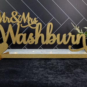 Op de afbeelding: Een gouden glitter bruiloft bord met de tekst "Mr. & Mrs. Washburn" en een hart. Het bord staat op een witte basis met lampjes.