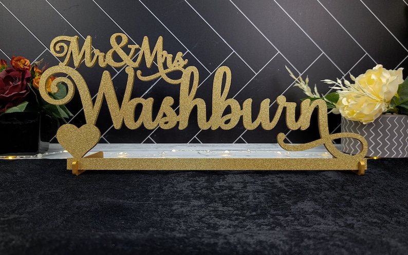 Op de afbeelding: Een gouden glitter bruiloft bord met de tekst "Mr. & Mrs. Washburn" en een hart. Het bord staat op een witte basis met lampjes.