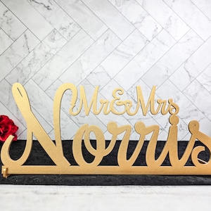 May include: Gold-colored wooden sign with the text "Mr. & Mrs. Norris" in a cursive font. The sign is perfect for a wedding or anniversary celebration.
