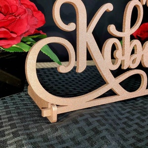 Op de afbeelding: Een roze gouden glitter bruiloft tafelbord met de namen "Mr. & Mrs. Kobasi" in een cursief schrift.