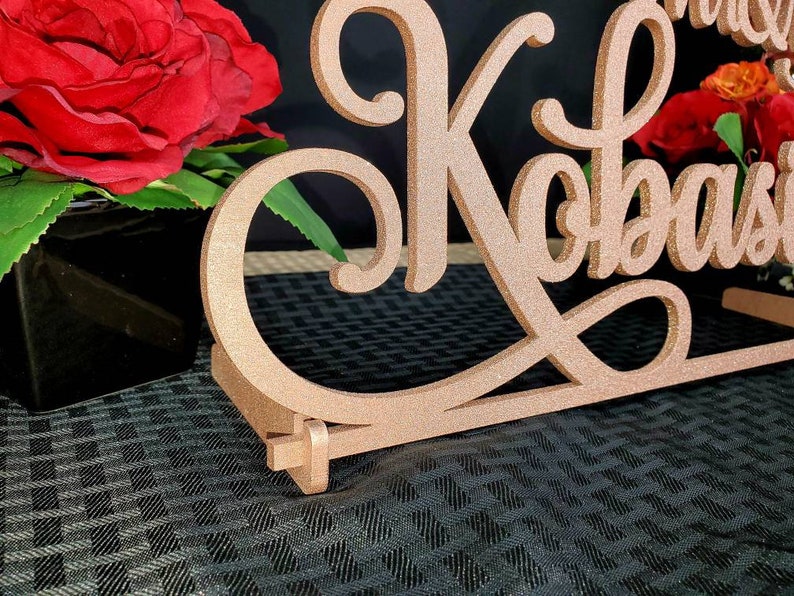 Op de afbeelding: Een roze gouden glitter bruiloft tafelbord met de namen "Mr. & Mrs. Kobasi" in een cursief schrift.