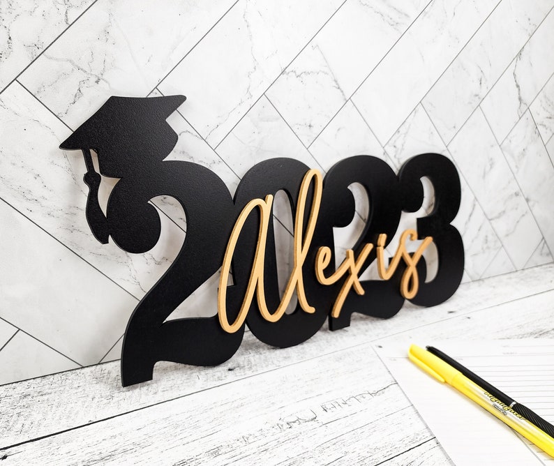 May include: Black wooden graduation sign with the year 2023 and the name Alexis in gold lettering. The number 2 is shaped like a graduation cap.