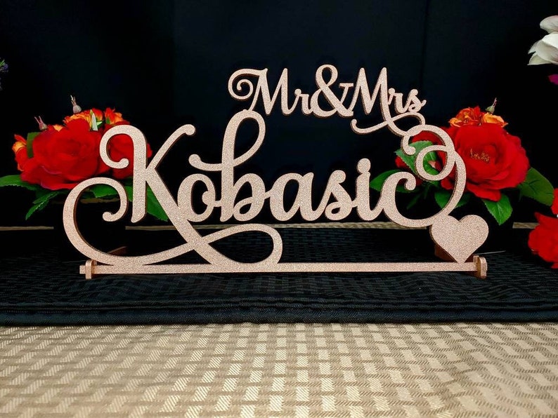 Op de afbeelding: Een roze gouden glitter bruiloft tafelbord met de tekst "Mr. & Mrs. Kobasic" in een decoratief handschrift. Een klein roze gouden hart is aan het bord bevestigd.