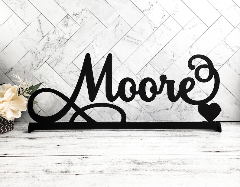 May include: Black metal sign with the word "Moore" in a cursive font with an infinity symbol and a heart. The sign is on a white wooden surface.