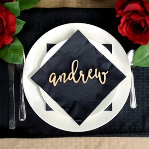 May include: A black napkin with a wooden name card that says "andrew" on a white plate. The plate is on a black tablecloth with red roses on either side.