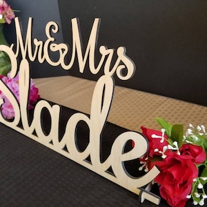 May include: Wooden wedding sign with the text "Mr. & Mrs. Slade" in cursive script. The sign is decorated with pink and red flowers.