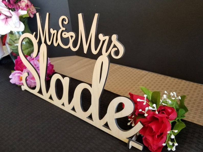 May include: Wooden wedding sign with the text "Mr. & Mrs. Slade" in cursive script. The sign is decorated with pink and red flowers.