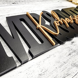 May include: Black wooden letters spelling "MR & MRS" with a gold cursive "MRS" overlay. The letters are layered and have a 3D effect.