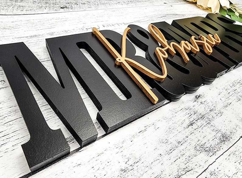 May include: Black wooden letters spelling "MR & MRS" with a gold cursive "MRS" overlay. The letters are layered and have a 3D effect.