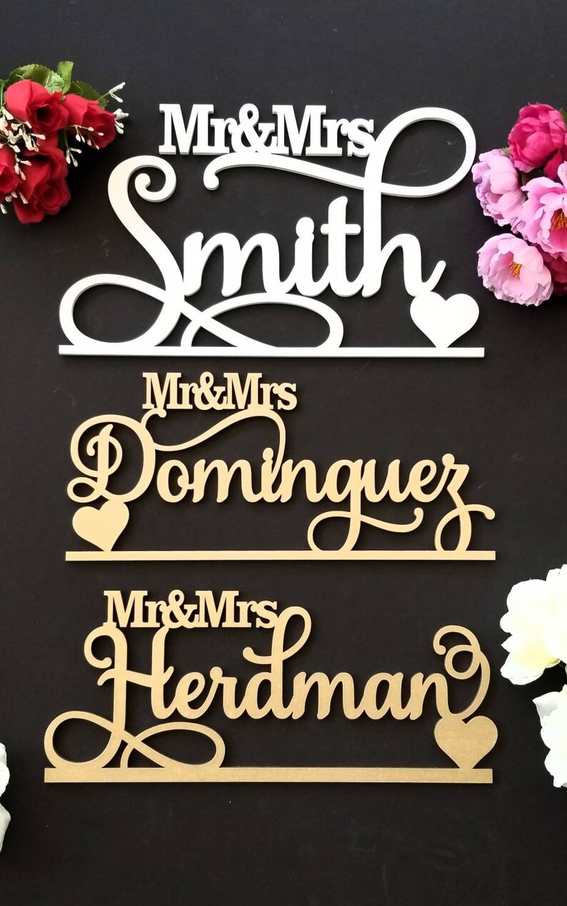 Op de afbeelding: Drie houten borden met de woorden "Mr. & Mrs. Smith", "Mr. & Mrs. Dominguez" en "Mr. & Mrs. Herdman" in een cursief schrift. Elk bord heeft een klein hartje aan het einde.