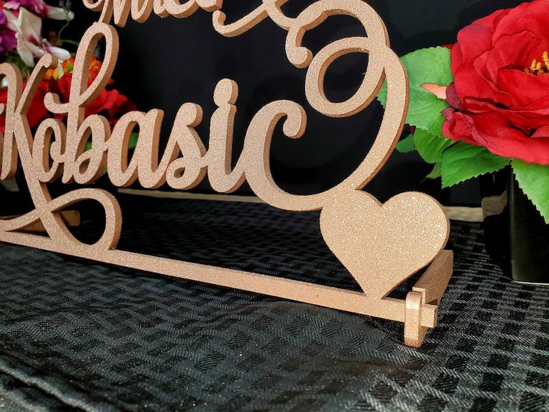 Op de afbeelding: Een houten bord met de woorden "Mr. & Mrs. Kobasic" in cursief schrift. Het bord is in een lichtbruine kleur geverfd en heeft een glitterafwerking. Een klein hartvormig uitsnijding is aan het bord bevestigd.