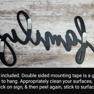 May include: Black metal wall decor with the word "julimah" written in a cursive script. Three white adhesive strips are attached to the back of the sign for hanging.