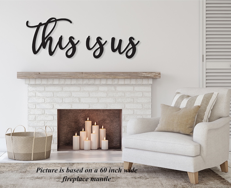 Op de afbeelding: Zwarte metalen wanddecoratie met de woorden "This is Us" in een cursief schrift. De decoratie is geplaatst boven een witte bakstenen open haard met een houten schouw. De open haard heeft kaarsen erin.