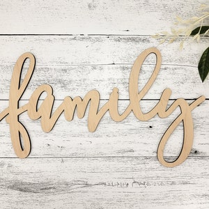 May include: Wooden sign with the word "family" written in cursive script. The sign is light brown and is placed on a white wooden background.