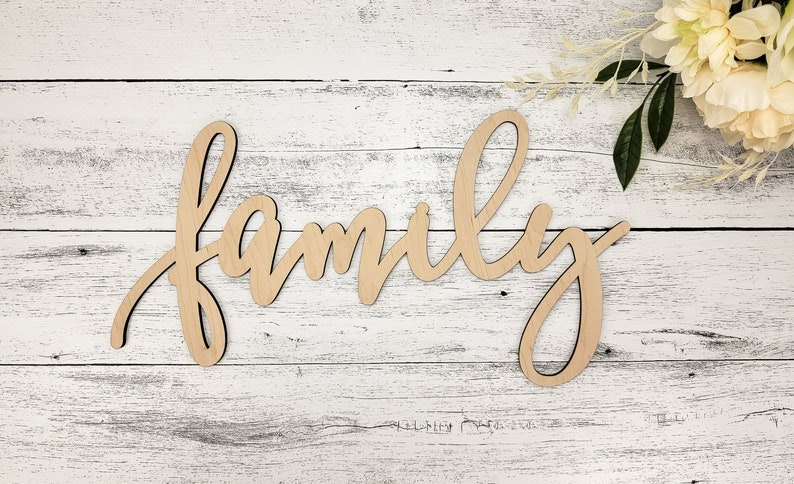 May include: Wooden sign with the word "family" written in cursive script. The sign is light brown and is placed on a white wooden background.