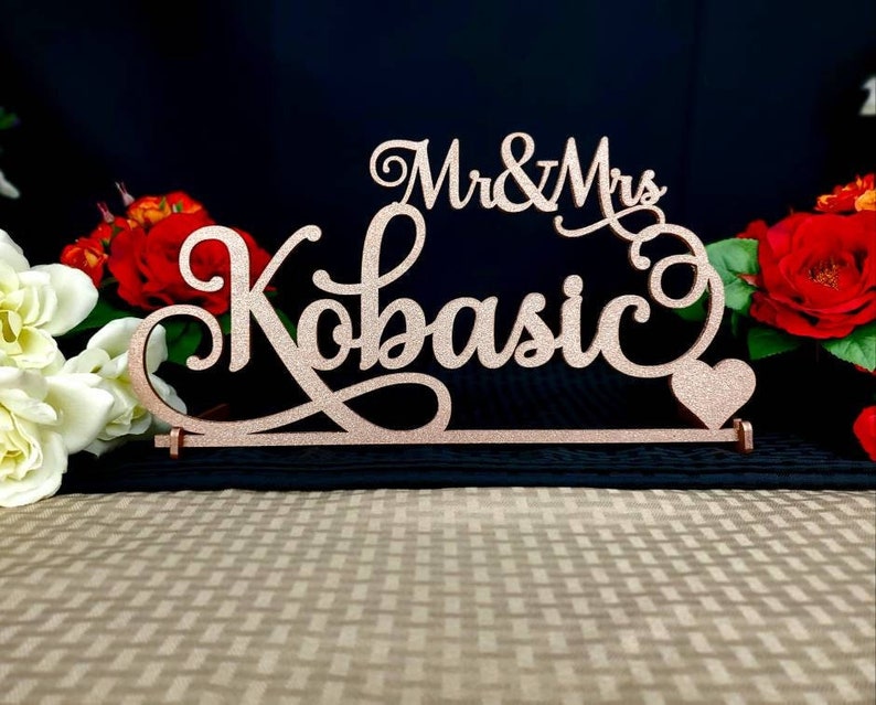 Op de afbeelding: Een roze gouden glitter bruidstaarttopper met de woorden "Mr & Mrs Kobasic" en een klein hartje.