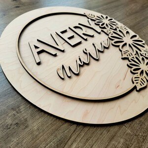 May include: A wooden round sign with a circle cut out in the center. The circle has the names "Avery" and "marie" written in a decorative font. The sign is decorated with a floral design around the edge.