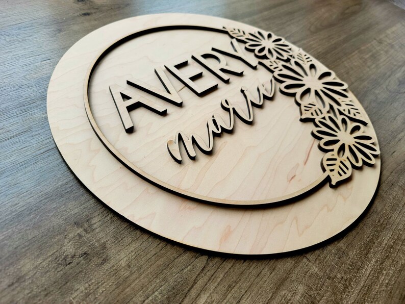 May include: A wooden round sign with a circle cut out in the center. The circle has the names "Avery" and "marie" written in a decorative font. The sign is decorated with a floral design around the edge.