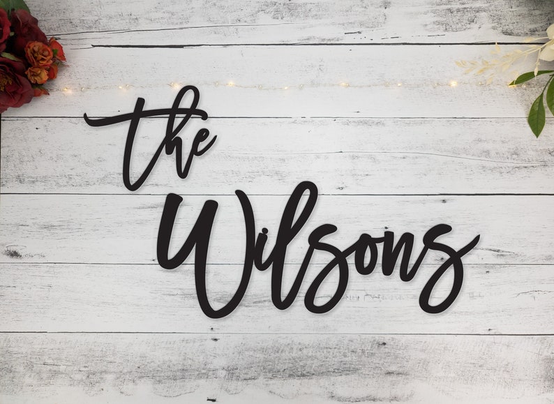 May include: Black wooden sign with the text "the Wilsons" in a cursive font on a white wood background.