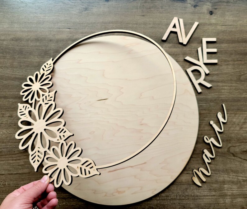 May include: A wooden craft kit for creating a personalized wreath. The kit includes a wooden circle with a cutout design, a floral design, and the words "AVRYE marie".