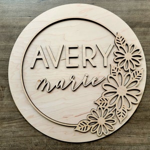 May include: A wooden round sign with a circle cut out in the center. The sign is decorated with a floral design and the names "Avery marie" are written in the center.