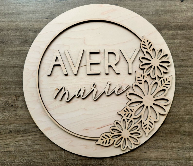 May include: A wooden round sign with a circle cut out in the center. The sign is decorated with a floral design and the names "Avery marie" are written in the center.
