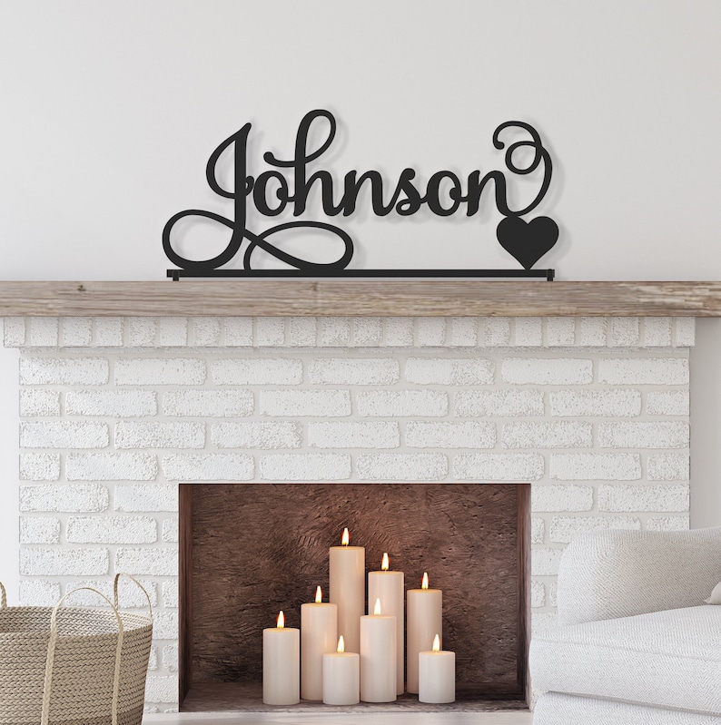 May include: Black metal sign with the name "Johnson" in a cursive font with a heart at the end. The sign is mounted above a white brick fireplace with candles inside.