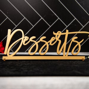 May include: A gold wooden sign with the word "Desserts" written in a cursive font. The sign is sitting on a black surface.