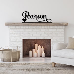 May include: Black metal sign with the name "Pearson" and a small heart above it, mounted on a white brick fireplace mantel. The fireplace has a wood-burning insert with lit candles inside.
