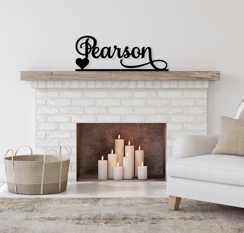 May include: Black metal sign with the name "Pearson" and a small heart above it, mounted on a white brick fireplace mantel. The fireplace has a wood-burning insert with lit candles inside.