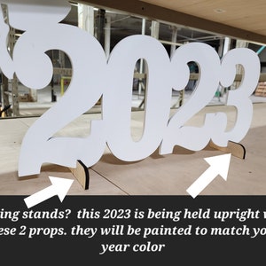 May include: White wooden 2023 cutout with two small wooden stands to hold it upright. The stands are painted to match the year color.
