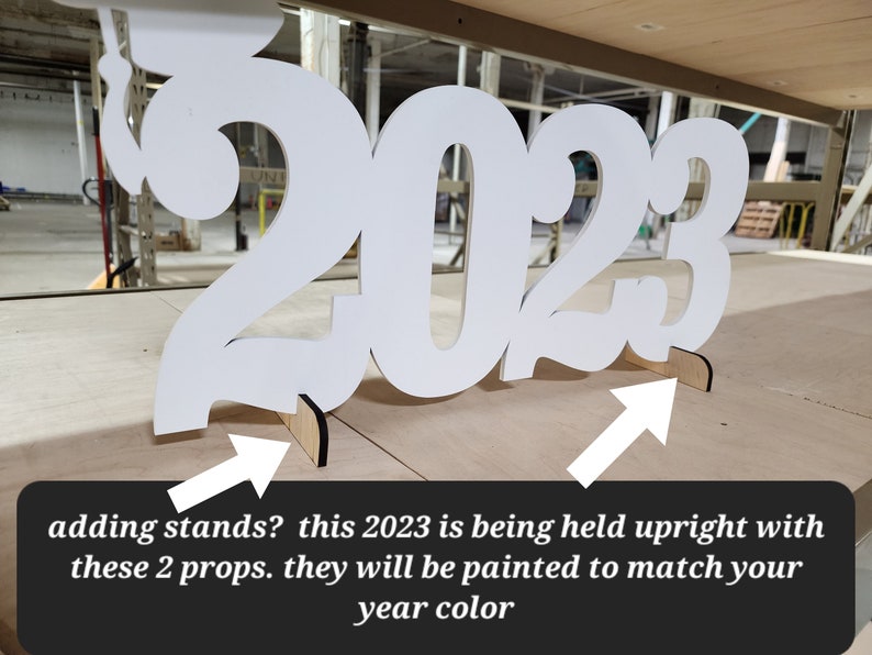 May include: White wooden 2023 cutout with two small wooden stands to hold it upright. The stands are painted to match the year color.
