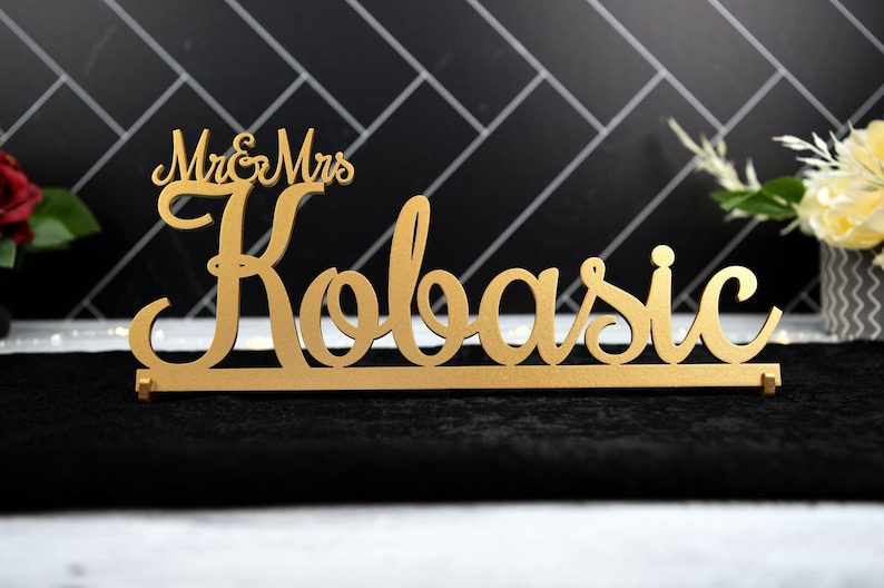 May include: Gold-colored wooden sign with the text "Mr. & Mrs. Kobasic" in a cursive font.