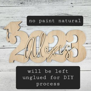 May include: Unpainted wooden cutout of the year 2023 with the name Alexis in cursive script. The cutout is designed for a graduation celebration. The text "no paint natural" and "will be left unglued for DIY process" is included in the image.