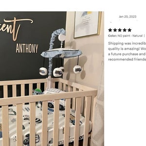 May include: A wooden crib with a white and blue truck themed bedding set. A wooden mobile with blue fabric and truck-shaped toys hangs above the crib. The crib is in front of a dark gray wall with a wooden sign that reads "Vincent" and "Anthony".