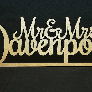 May include: Gold glitter Mr. & Mrs. Davenport wedding cake topper. The topper is made of wood and features a heart shape at the end.