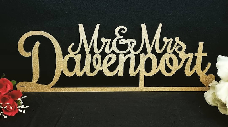 May include: Gold glitter Mr. & Mrs. Davenport wedding cake topper. The topper is made of wood and features a heart shape at the end.