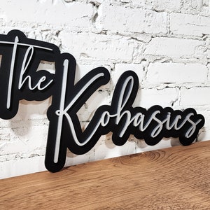 May include: Black wooden sign with the text "The Kohasics" in white lettering. The sign is in a cursive font and has a 3D effect.