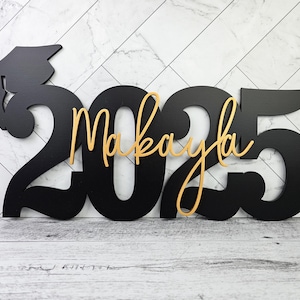 May include: Black wooden cutout of the year 2025 with a graduation cap and the name Makayla in gold lettering.