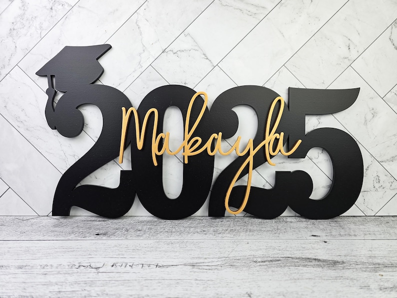 May include: Black wooden cutout of the year 2025 with a graduation cap and the name Makayla in gold lettering.