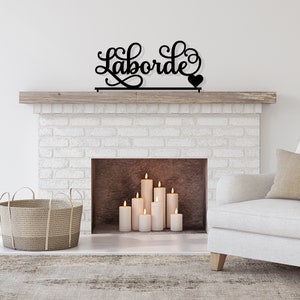 May include: Black metal sign with the word "Laborde" in a cursive font with a small heart at the end. The sign is mounted above a white brick fireplace with a wooden mantel. There are several white candles in the fireplace.