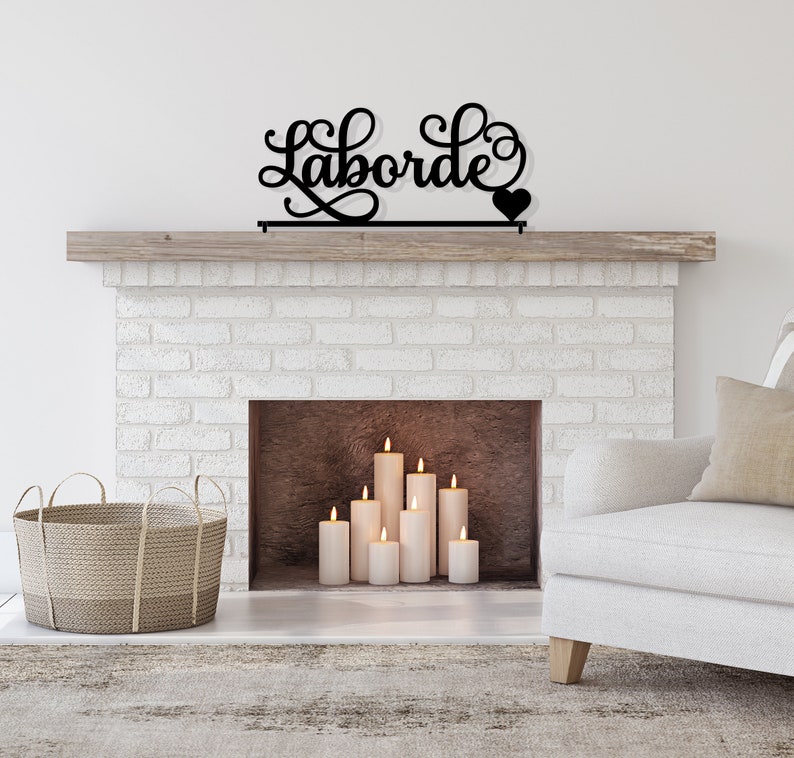 May include: Black metal sign with the word "Laborde" in a cursive font with a small heart at the end. The sign is mounted above a white brick fireplace with a wooden mantel. There are several white candles in the fireplace.