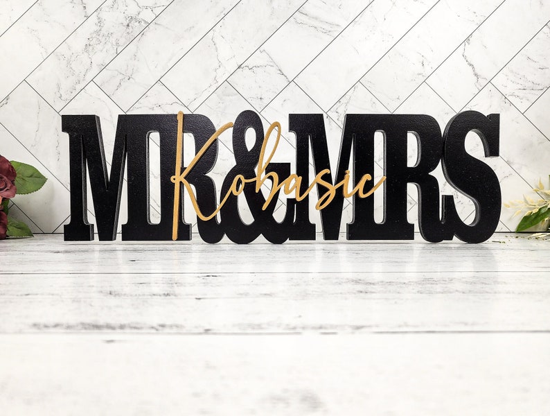 May include: Black wooden "Mr. & Mrs." sign with a gold cursive "Kobasic" in the center. The sign is on a white wooden surface.