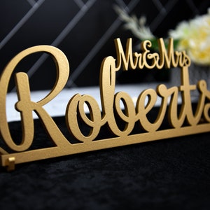 May include: Gold-colored wooden sign with the text "Mr. & Mrs. Roberts" in a cursive font.