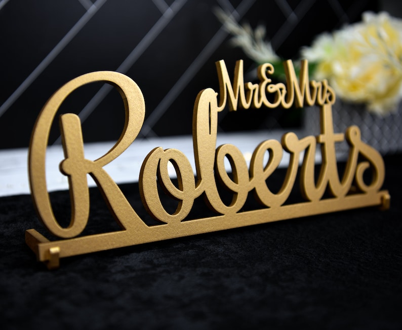 May include: Gold-colored wooden sign with the text "Mr. & Mrs. Roberts" in a cursive font.