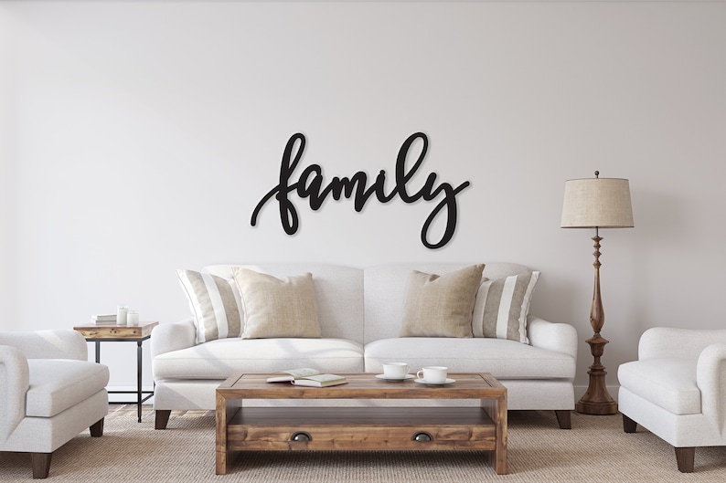 May include: Black wooden wall decor featuring the word "family" in a cursive font. The decor is displayed above a white sofa with cushions in a living room setting.