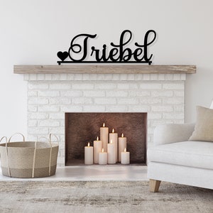 May include: A black metal sign with the word "Trieber" in a cursive font, with a small heart on the left side of the word. The sign is mounted above a white brick fireplace with a wooden mantel. There are several white candles in the fireplace.