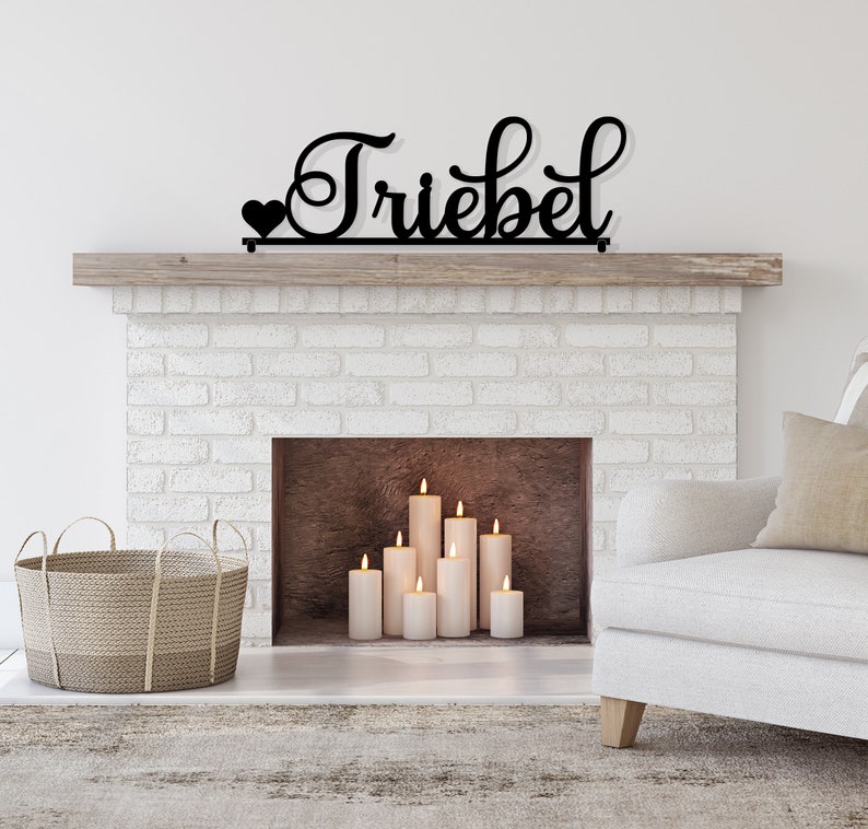 May include: A black metal sign with the word "Trieber" in a cursive font, with a small heart on the left side of the word. The sign is mounted above a white brick fireplace with a wooden mantel. There are several white candles in the fireplace.
