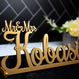 May include: Gold-colored wooden sign with the text "Mr. & Mrs. Kobasic" in a cursive font.